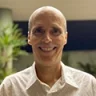 unknown: Retrato em plano médio de um homem sorridente de meia-idade, careca, vestindo uma camisa social branca.