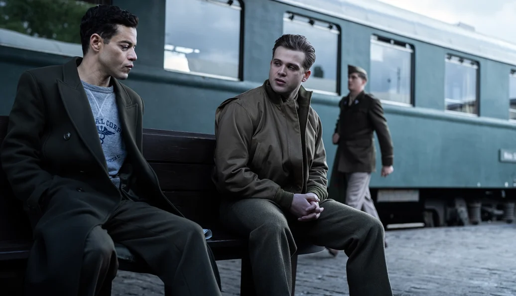 Rami Malek, Josh Peck: Still do filme Oppenheimer mostrando Rami Malek e Josh Peck conversando sentados em um banco em frente a um trem.