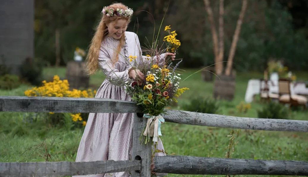 Eliza Scanlen: Uma mulher ruiva com coroa de flores e vestido de época segurando um buquê de flores silvestres junto a uma cerca de madeira.