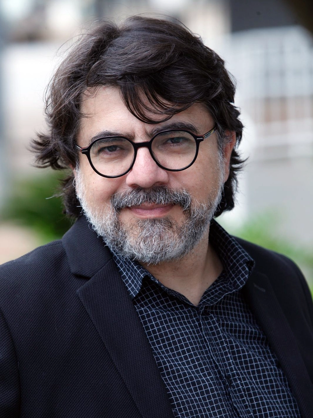 Javier Cercas: Retrato de um homem com óculos redondos, barba grisalha e cabelo escuro vestindo um blazer preto sobre camisa quadriculada.