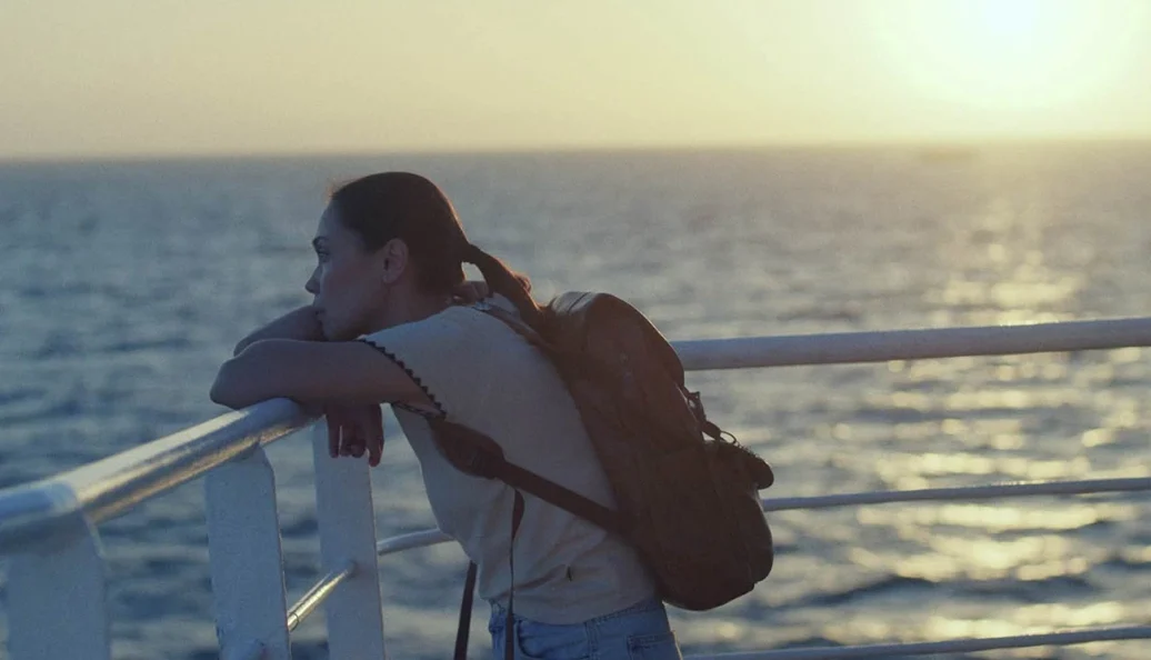 desconhecido: Uma mulher de mochila encostada no corrimão de um navio observando o mar durante o pôr do sol.
