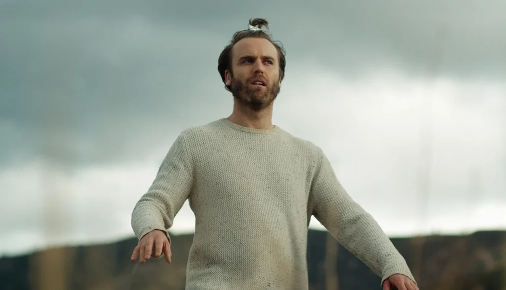Joshua Brennan: Homem com coque no cabelo e barba, vestindo uma camisola bege em um ambiente externo sob céu nublado.