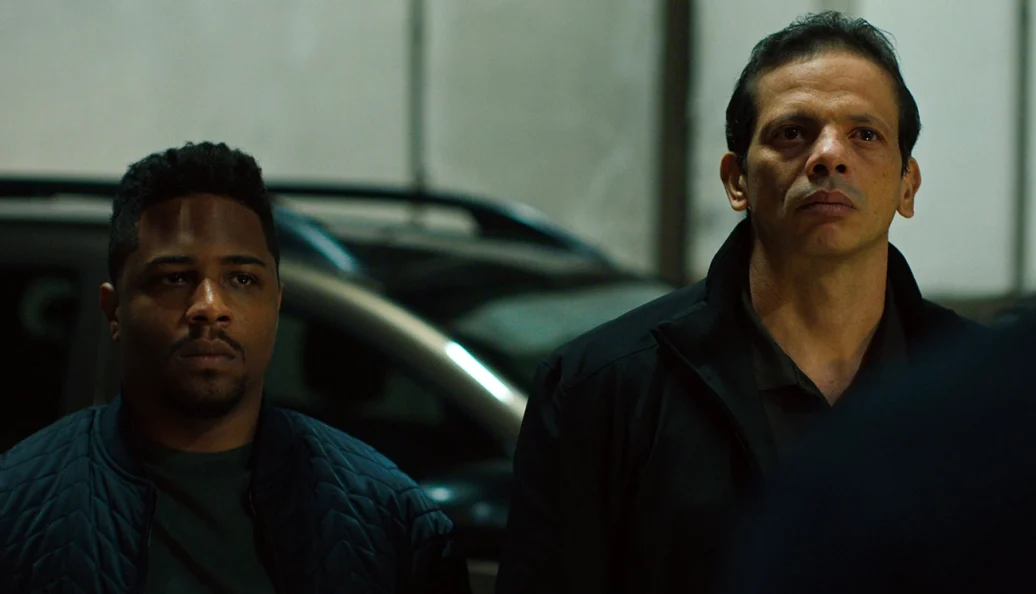 Digão Ribeiro e Ricardo Gelli: Dois homens com expressões sérias em um ambiente que parece ser um estacionamento, cena do filme Alemão 2.