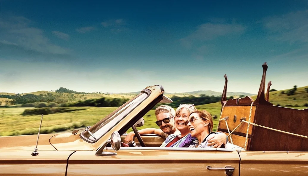 unknown: Uma cena de filme mostrando três pessoas sorridentes em um carro conversível carregado com móveis, viajando por uma estrada rural.
