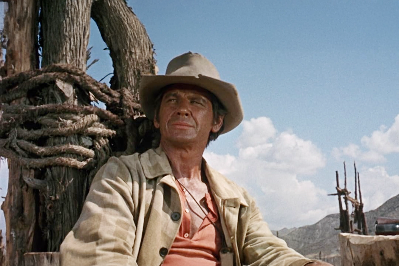 Charles Bronson: Cena do filme Era Uma Vez no Oeste (Once Upon a Time in the West) com um cowboy sentado sob o sol.