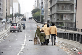 Uma cena de filme mostrando pessoas caminhando por uma via urbana deserta e degradada com carros abandonados, remetendo a um cenário pós-apocalíptico.