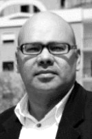 João Tordo: Retrato em preto e branco de um homem calvo usando óculos, camisa branca e paletó.