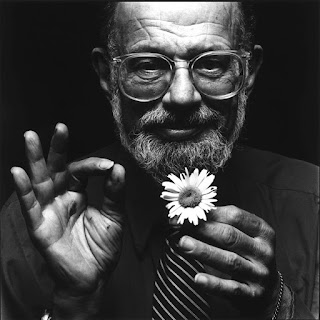 Allen Ginsberg: Retrato em preto e branco de um homem barbudo com óculos, segurando uma pequena flor e fazendo um gesto de 'ok' com a mão.