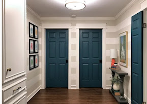 unknown: Interior de um corredor decorado com duas portas azuis, paredes listradas em branco e cinza, quadros na parede esquerda e um pequeno aparador na direita.