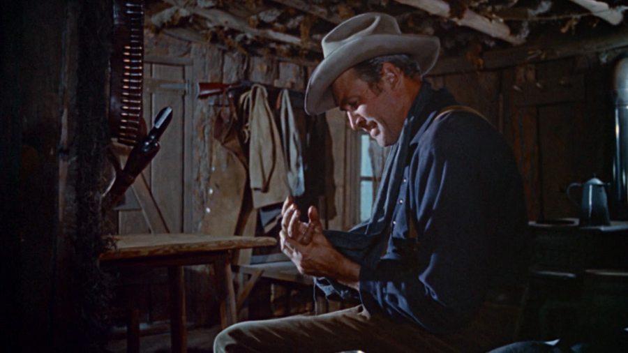 James Stewart: Cena de um filme de faroeste onde um homem sentado em uma cabana rústica observa suas mãos feridas.