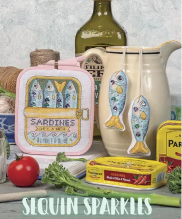 A still life arrangement featuring cross-stitch embroidery of sardine tins and fish, decorated with sequins, alongside real kitchen items like olive oil and vegetables.