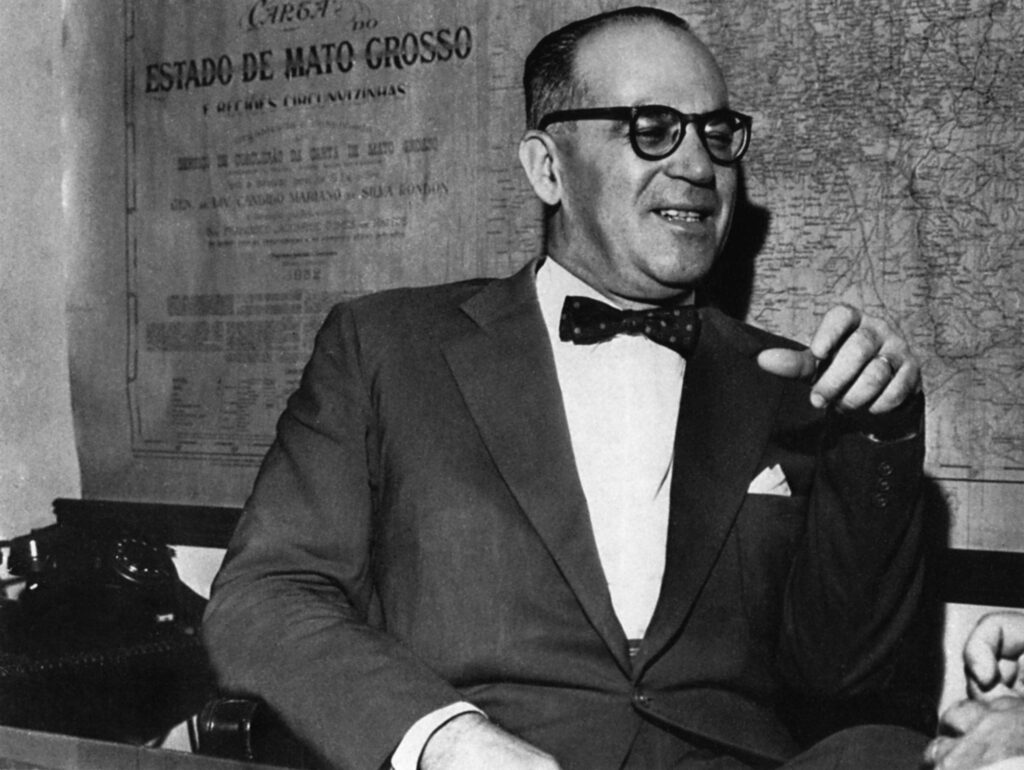 João Guimarães Rosa: Retrato em preto e branco de um homem sorridente usando óculos, terno, gravata borboleta e um lenço no bolso, sentado em frente a um mapa do Estado de Mato Grosso.