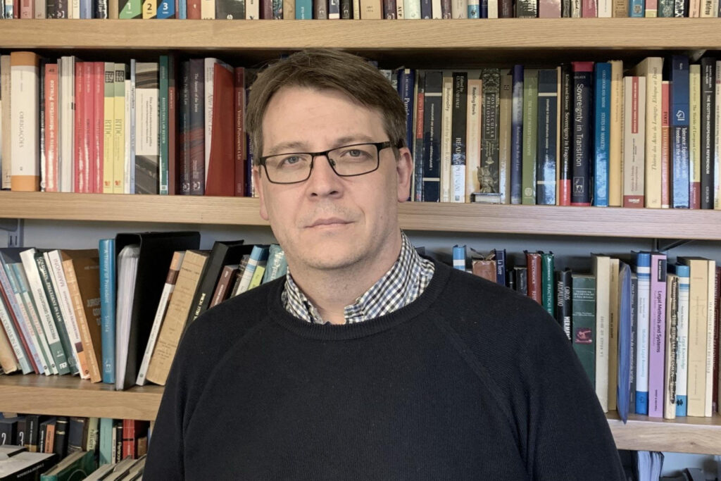 Nuno Garoupa: Retrato de um homem usando óculos e suéter escuro em frente a uma estante repleta de livros.