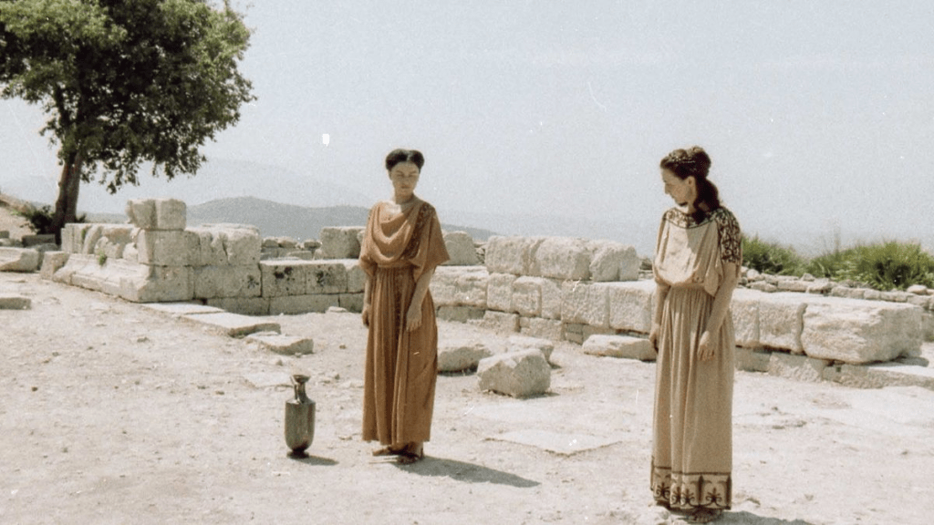 unknown: Duas mulheres vestidas com túnicas de estilo grego antigo em pé entre ruínas de pedra ao ar livre.