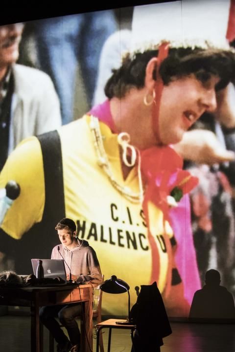 Um homem sentado à frente de um laptop em um palco com uma grande projeção ao fundo mostrando uma pessoa com traje amarelo e chapéu branco.