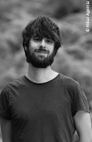 unknown: Retrato em preto e branco de um homem jovem com barba e cabelo escuro, vestindo uma camiseta escura.