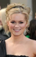 Alice Eve: Retrato de uma mulher loira sorrindo, usando uma tiara preta e um vestido de um ombro só.