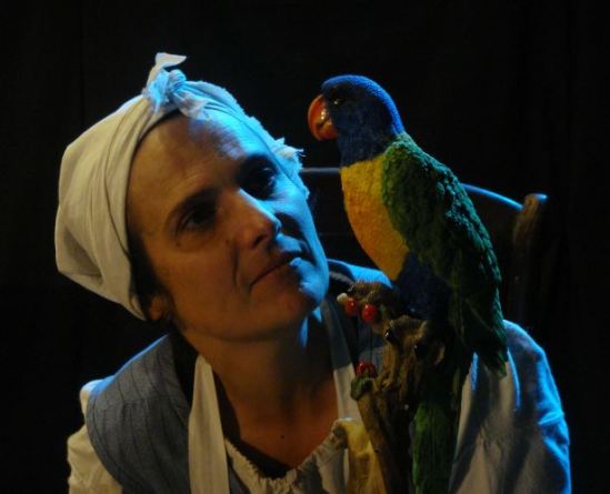 Uma mulher usando um lenço branco na cabeça olha para um papagaio colorido artificial em um ambiente escuro, possivelmente uma cena teatral.