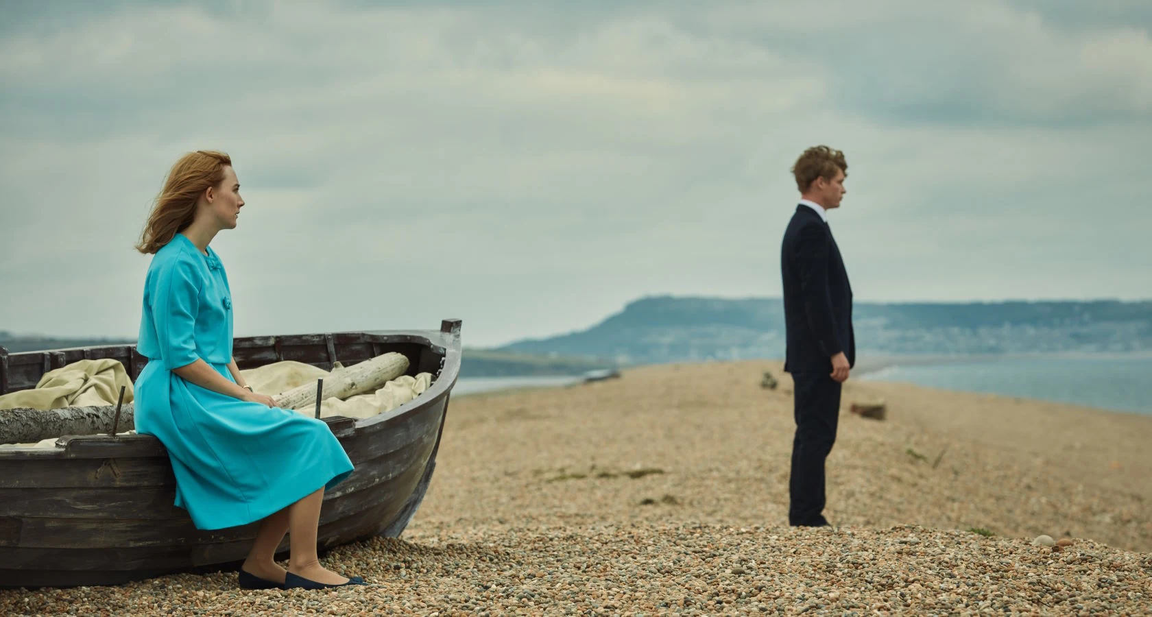 On Chesil Beach Saoirse Ronan, Billy Howle: Uma mulher vestida de azul sentada em um barco em uma praia de seixos, enquanto um homem de terno está de pé ao fundo, ambos olhando em direções opostas sob um céu nublado.
