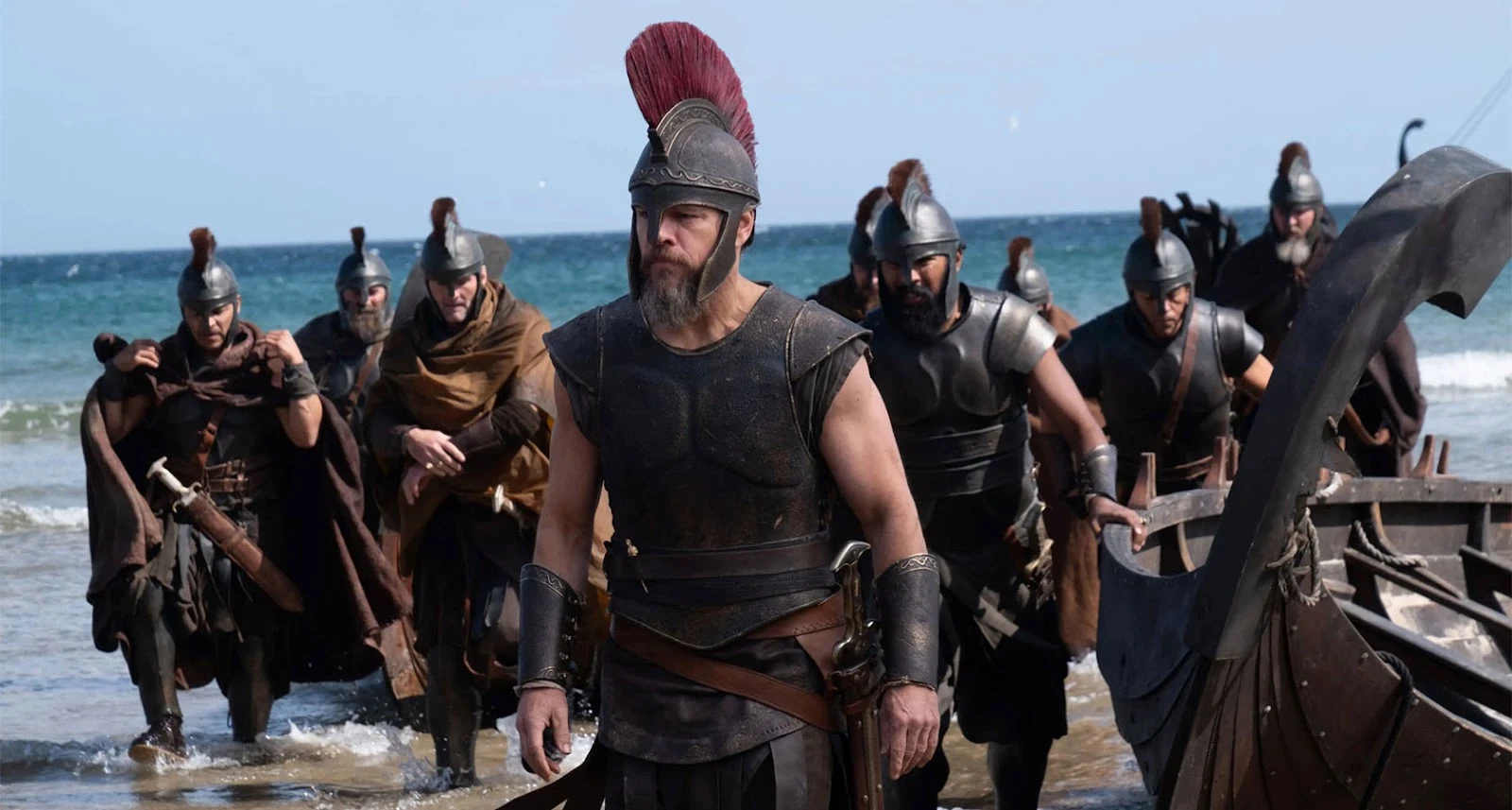 unknown: Cena de filme ou série mostrando guerreiros gregos ou romanos desembarcando em uma praia com armaduras e elmos antigos.