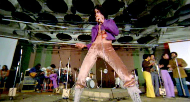 Caetano Veloso: Performancce de música ao vivo com cantor em destaque no centro do palco usando jaqueta roxa aberta e calças de veludo.