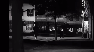 desconhecido: Uma cena de filme em preto e branco mostrando uma rua de cidade à noite com uma figura solitária caminhando.