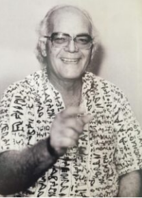 Glauber Rocha: Retrato em preto e branco de um homem sorridente usando óculos e uma camisa estampada, apontando para a câmera.