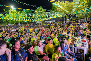 Uma grande multidão de pessoas vestidas com as cores do Brasil (amarelo, verde e azul) assistindo a um evento ao ar livre à noite, com bandeirinhas decorativas sobre o público.