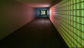 Um corredor longo e escuro com uma parede lateral feita de blocos de vidro iluminados em tons de verde e vermelho.