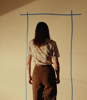 desconhecido: Uma mulher de costas parada em frente a uma parede onde há o desenho azul de um retângulo ou porta.
