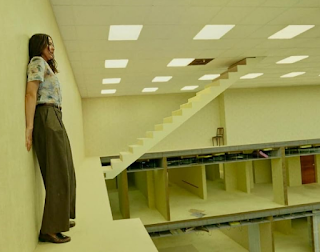 unknown: Uma mulher encostada em uma parede em um cenário surrealista que lembra o conceito de 'Backrooms', com escadas que levam ao teto e múltiplos níveis de salas vazias de escritório.