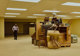 unknown: A man walking past a pile of furniture in a large room with yellow walls and fluorescent lighting, resembling 'The Backrooms'.