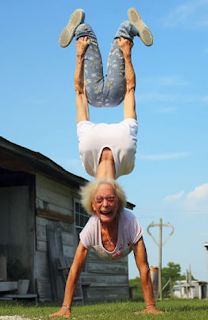 unknown: Uma mulher idosa fazendo uma parada de mão com outra pessoa equilibrada sobre seus ombros, criando uma ilusão de ótica.