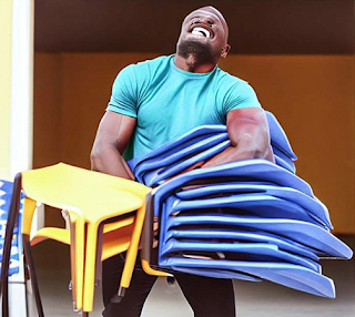 desconhecido: Homem musculoso carregando uma grande pilha de cadeiras plásticas azuis e amarelas.