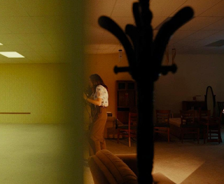 desconhecido: Uma cena cinematográfica mostrando uma mulher em um ambiente interno mal iluminado com móveis, vista através de uma obstrução em primeiro plano.