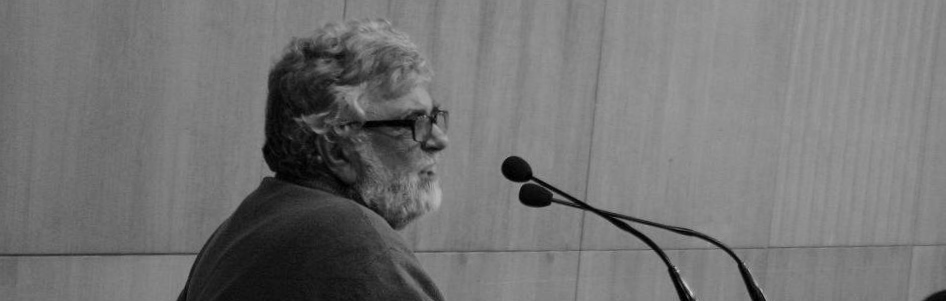 Luis Sepúlveda: Fotografia em preto e branco de um homem idoso com barba e óculos falando ao microfone.