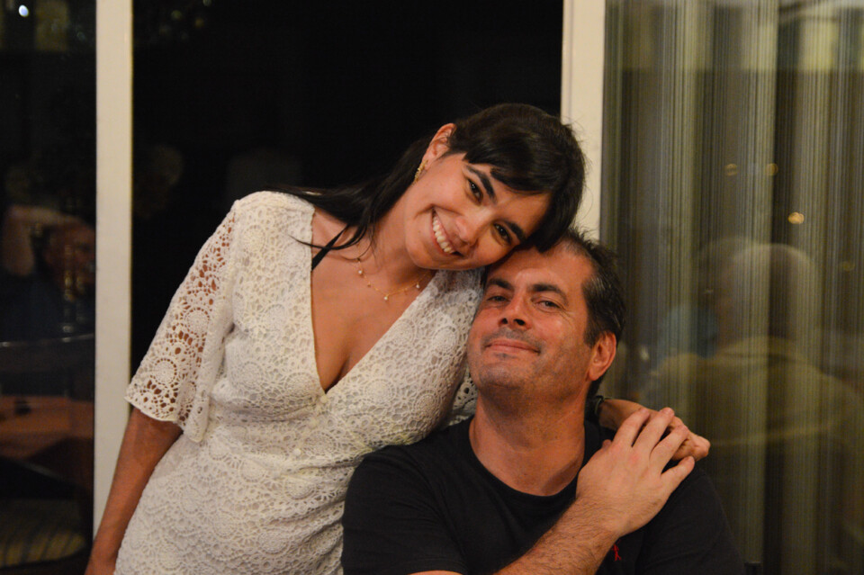 Uma mulher de vestido branco rendado sorrindo e encostando a cabeça em um homem de camiseta preta.