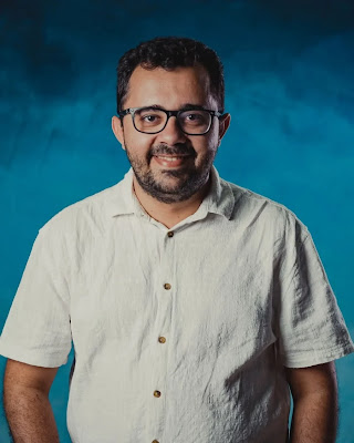 unknown: Retrato de um homem sorridente com óculos e camisa branca de botões em frente a um fundo azul.