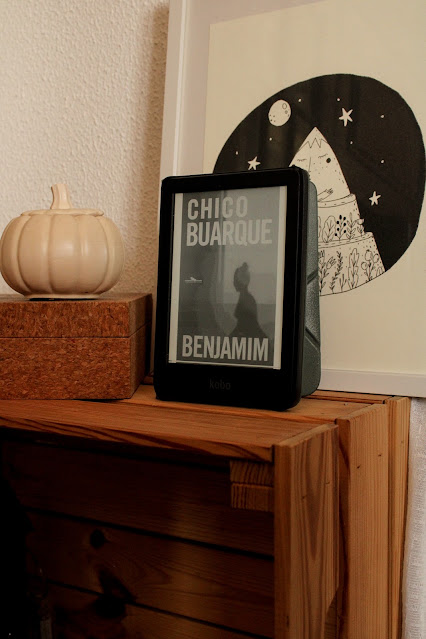 Chico Buarque: Um e-reader Kobo exibindo a capa do livro Benjamim de Chico Buarque, posicionado sobre um móvel de madeira ao lado de uma abóbora decorativa e um quadro.