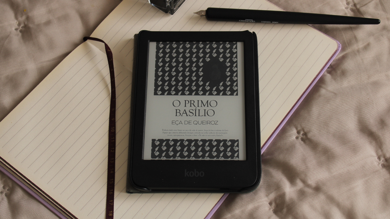 Um e-reader Kobo exibindo a capa do livro 'O Primo Basílio' de Eça de Queiroz, posicionado sobre um caderno aberto ao lado de uma caneta tinteiro.
