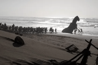 Uma fotografia em preto e branco de uma cena de filme mostrando um grande grupo de pessoas em uma praia puxando uma estátua gigante ou estrutura em forma de cavalo para fora da água.