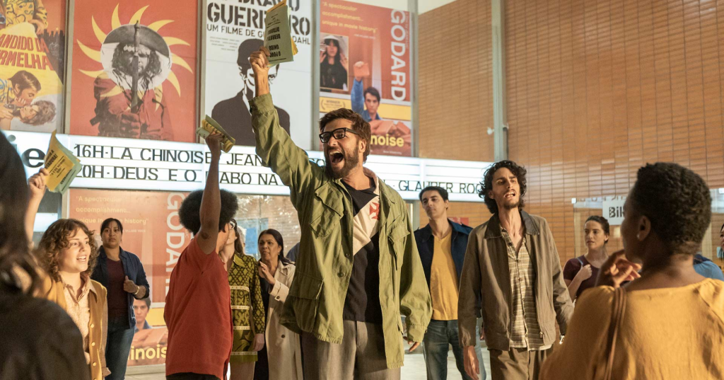 Bruno Gagliasso: Uma cena do filme 'O Paciente Interno' ou similar, mostrando um grupo de pessoas em um cinema antigo com cartazes de filmes de Glauber Rocha e Godard ao fundo, com um homem central gritando e levantando panfletos.