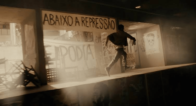 unknown: Uma cena cinematográfica em tons sépia mostrando um homem correndo em um ambiente urbano com cartazes de protesto, incluindo um que diz 'ABAIXO A REPRESSÃO'.
