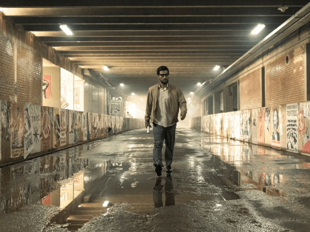 Lázaro Ramos: Um homem caminha por um túnel urbano úmido e iluminado, com cartazes colados nas paredes laterais.