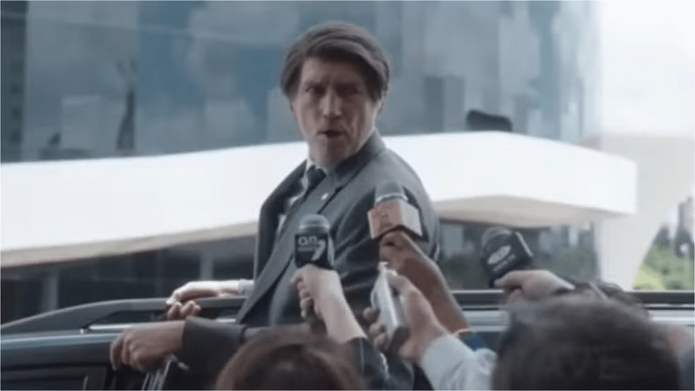 Ator interpretando personagem semelhante a Jair Bolsonaro: Homem de terno, assemelhando-se a um político, cercado por microfones de repórteres em uma coletiva de imprensa ao ar livre.