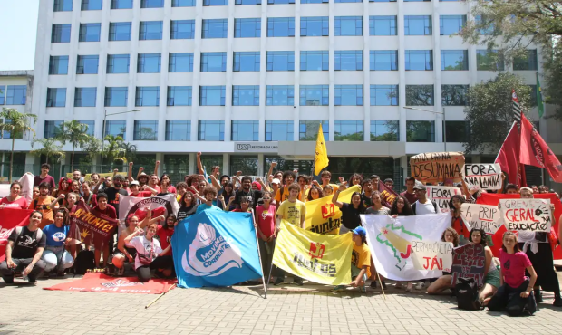 Estudantes em protesto em frente ao prédio da Reitoria da USP, segurando bandeiras e cartazes reivindicando permanência estudantil e outras pautas.