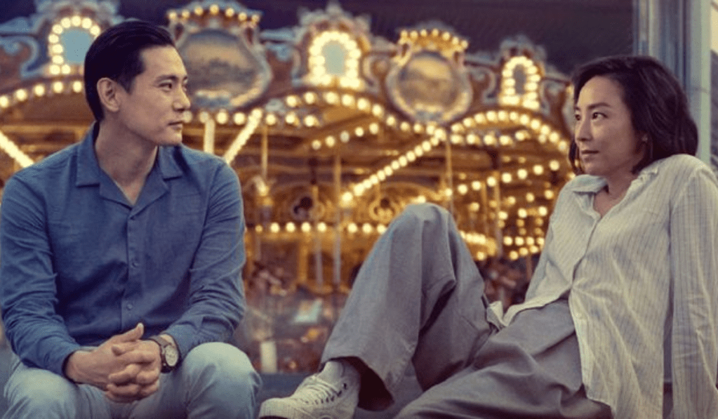 Teo Yoo, Greta Lee: Uma cena do filme Past Lives mostrando um homem e uma mulher sentados em frente a um carrossel iluminado à noite.