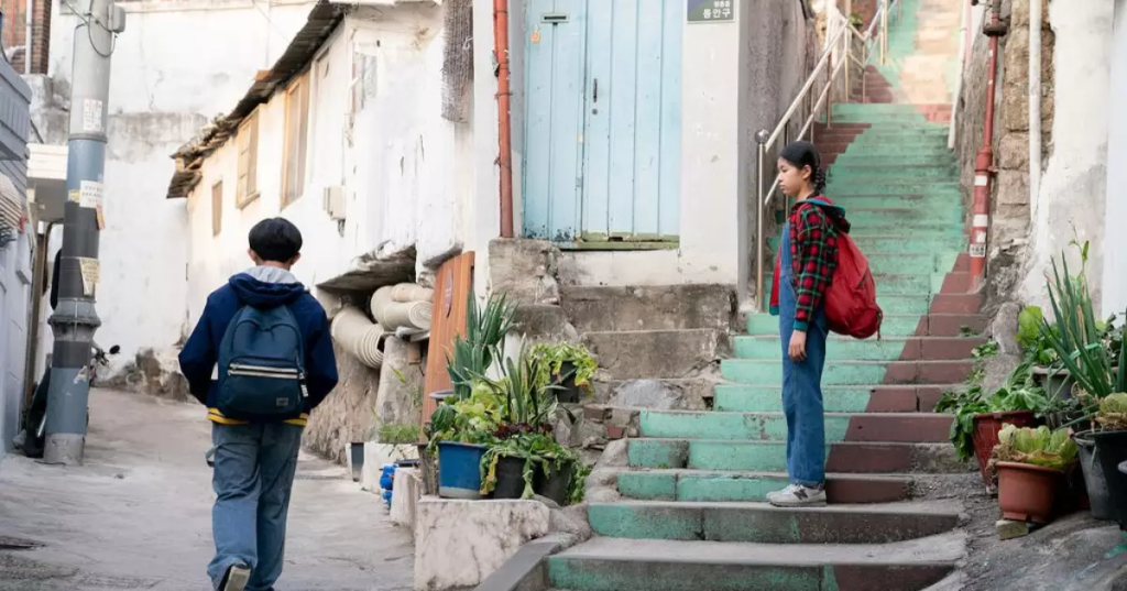 Cena de filme mostrando um menino de costas caminhando em um beco estreito em direção a uma menina que está parada em uma escadaria colorida.