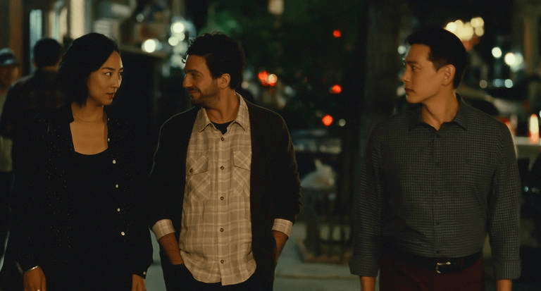Greta Lee, John Magaro, Teo Yoo: Uma cena do filme Past Lives mostrando três personagens caminhando à noite em uma rua urbana.