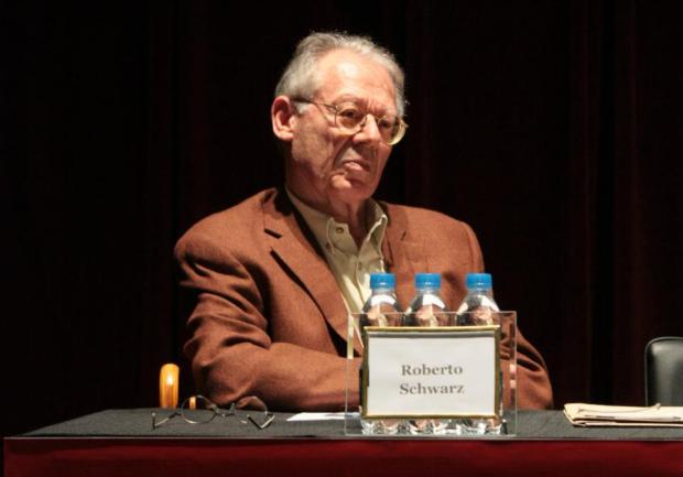 Roberto Schwarz: Homem idoso de óculos e paletó marrom sentado à mesa em um evento, com uma placa de identificação à sua frente.